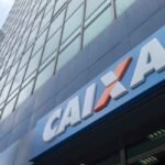 Fachada de uma agência da Caixa Econômica Federal em um edifício comercial de vidro. Em primeiro plano, uma placa azul exibe o logotipo e o nome “CAIXA” em letras brancas com detalhe laranja. Ao fundo, a torre do prédio se eleva com inúmeras janelas refletindo o céu, enquanto outro edifício alto aparece parcialmente à esquerda. A imagem foi capturada de baixo para cima, destacando a arquitetura urbana e a sinalização da instituição financeira.