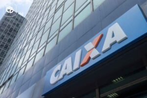 Fachada de uma agência da Caixa Econômica Federal em um edifício comercial de vidro. Em primeiro plano, uma placa azul exibe o logotipo e o nome “CAIXA” em letras brancas com detalhe laranja. Ao fundo, a torre do prédio se eleva com inúmeras janelas refletindo o céu, enquanto outro edifício alto aparece parcialmente à esquerda. A imagem foi capturada de baixo para cima, destacando a arquitetura urbana e a sinalização da instituição financeira.